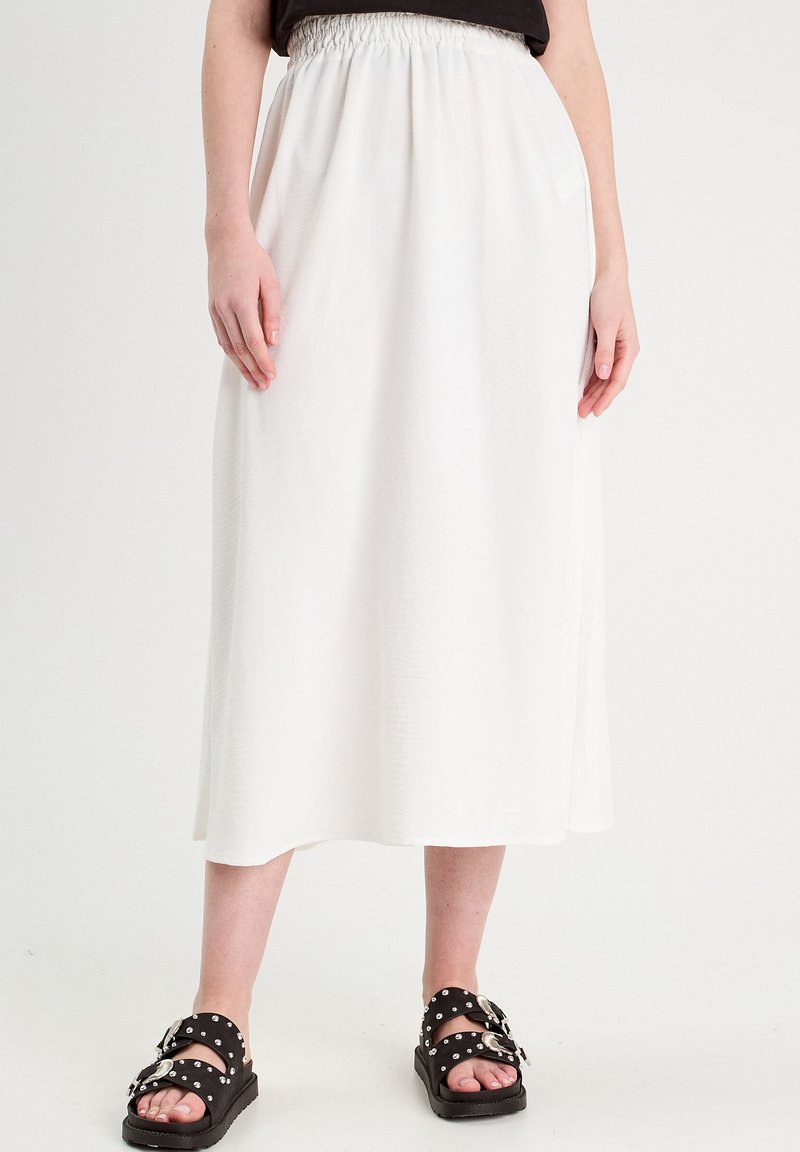 Jupe midi blanche avec une taille élastique, présentant une texture douce et un design fluide, associée à des sandales noires ornées de clous en argent.