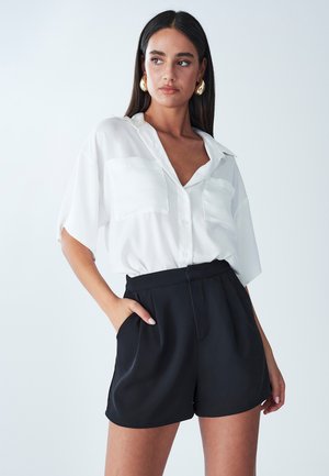 Camicia bianca con bottoni e maniche corte, dotata di due tasche sul petto, abbinata a pantaloni corti neri a vita alta. Tessuto morbido, vestibilità casual.