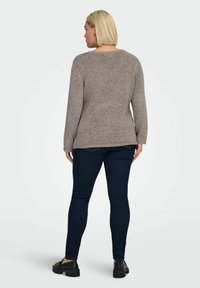 Žena s krátkými blond vlasy, oblečená v béžovém pleteném svetru, tmavých úzkých džínách a černých masivních mokasínách, stojící zády ke kameře.