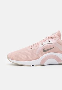 Nike-kengät, joissa on vaaleanpunainen verkko-olka, pyöreä kärki ja harmaat yksityiskohdat. Ominaisuuksiin kuuluu teksturoitu muotoilu sekä valkoinen välipohja, jossa on kaareva pohjamuoto.