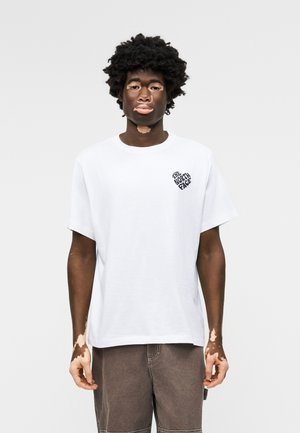 Νεαρός άνδρας με λεύκη που φοράει λευκό μπλουζάκι The North Face και καφέ παντελόνι, στέκεται μπροστά σε ένα απλό ανοιχτό γκρι φόντο.