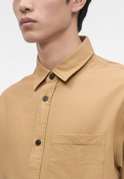 Gros plan d’un homme portant une chemise marron clair texturée boutonnée avec un col et une poche avant sur fond blanc.