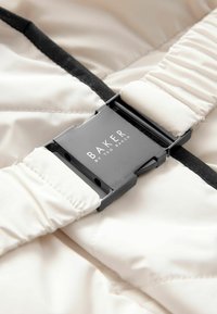 Cremefarbene, gesteppten Stoff mit einem schwarzen Riemen und einer silbernen Schnalle. Die Schnalle ist mit "BAKER by TED BAKER" graviert.