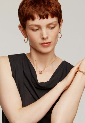 Collarín en forma de estrella dorada con acentos de piedras preciosas rosas, llevado sobre una blusa oscura sin mangas. Pendientes de aro dorados simples complementan el look.