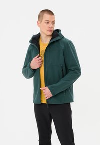 Veste softshell vert foncé avec un devant zippé, une capuche et deux poches latérales. Portée sur un t-shirt jaune, présentant un design graphique subtil.