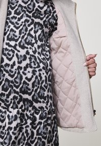 Rankomis laikomas atidarytas smėlio spalvos paltas su išsiuvinėta šviesiai rožine pamušalu, po juo matosi juodos ir pilkos leopardo rašto suknelė.