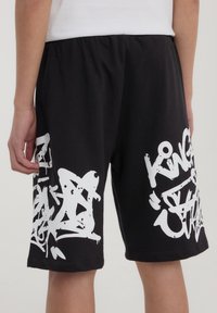 Pantaloni corti neri in cotone con ampio testo e design in stile graffiti bianchi sui lati inferiori. Vita elastica per il massimo comfort.