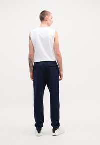 Camisa sin mangas blanca y pantalones azul marino. Los pantalones tienen un bolsillo trasero. La modelo está de espaldas, mostrando el conjunto y el ajuste.