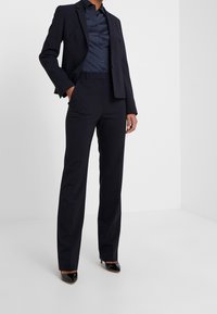 Costume sur mesure bleu marine foncé avec un blazer ajusté et un pantalon droit. Tissu lisse avec un léger éclat, associé à des escarpins noirs pointus.