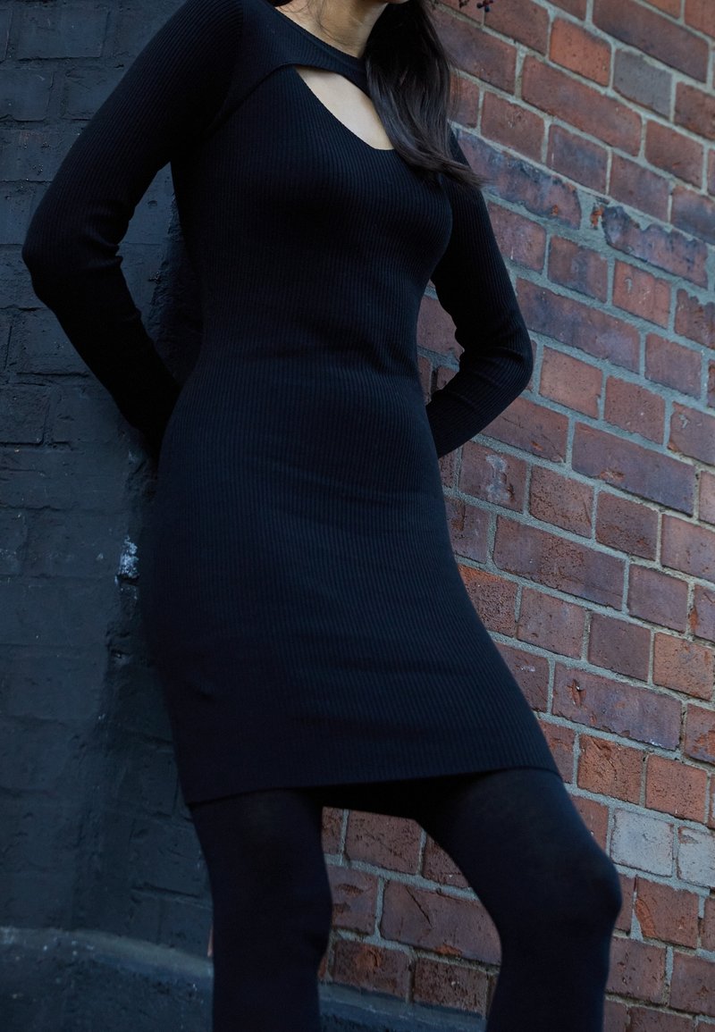 Robe noire côtelée avec un décolleté en forme de cœur et un détail découpé. Manches longues, silhouette ajustée et tissu texturé contre un mur en briques.