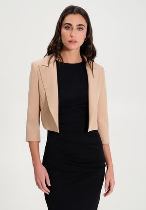Beige cropped blazer met ingesneden revers, driekwart mouwen en gladde stof, gedragen over een aansluitende zwarte jurk.