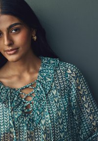 Blouse bleue à motifs avec un col froncé et un détail lacé. Tissu lisse présentant des motifs de cœurs et de feuilles. Boucles d'oreilles en forme de créole dorées.