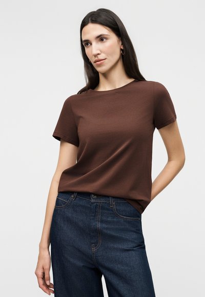 Γυναίκα με μακριά σκουρόχρωμα μαλλιά, ντυμένη με καφέ κοντομάνικο πουκάμισο και σκούρα μπλε τζιν, στέκεται μπροστά σε ένα απλό ανοιχτόχρωμο φόντο.