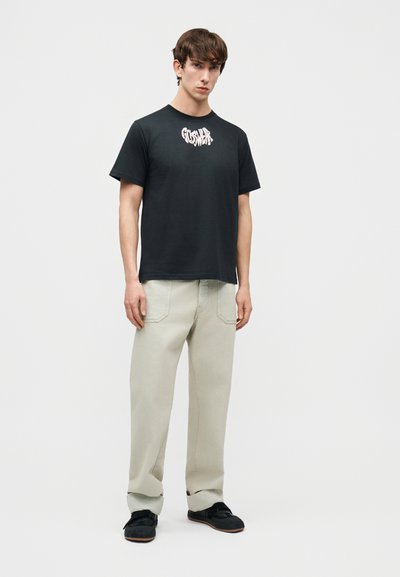 Νεαρός άνδρας στέκεται φορώντας μαύρο T-shirt με γραφικό σχέδιο, ανοιχτό μπεζ χαλαρό παντελόνι και μαύρα παπούτσια, μπροστά σε απλό φωτεινό φόντο.