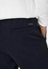 Marineblauwe broek van gladde stof, met een achterzak met een subtiel label. De broek heeft een klassieke pasvorm en een recht model.