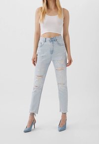 Ljusblå högmidjade jeans med slitna detaljer, som har fransade fållar och stora hål. Täckt med en vit kort crop-top och blå klackar.