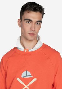 Sudadera naranja con cuello redondo, que presenta un gráfico de un sombrero y palas cruzadas. Llevada sobre una camisa de cuello claro. Textura suave.