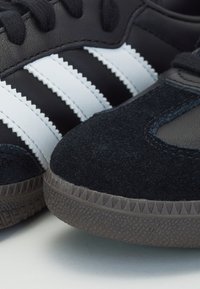 Basket en cuir noir avec des bandes blanches, bout en daim noir texturé et semelle en caoutchouc comportant des motifs triangulaires. Vue en gros plan.