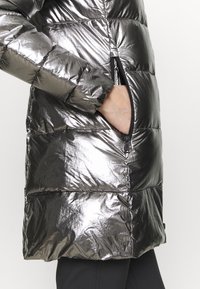 Doudoune métallique avec sections matelassées, poche latérale zippée et texture lisse et brillante. Couleur principalement gris argenté.