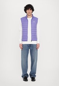 Colete puff estilo roxo com costura horizontal, fecho éclair na frente, gola alta, usado sobre uma camisa branca de mangas longas e jeans azuis.