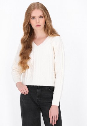 Kobieta z długimi falowanymi blond włosami, ubrana w biały sweter w warkocze i czarne jeansy na jednolitym białym tle.