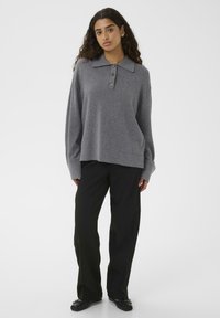 Woman with long curly hair wearing a loose gray buttoned sweater, black wide-leg pants, and black ballet flats on a white background.