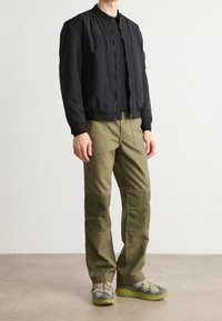 Chaqueta bomber negra, pantalones cargo verde oliva, zapatillas gris y verde. Telas ligeras y texturizadas con bolsillos funcionales y cremalleras.