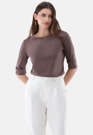 Frau mit langen braunen Haaren, die Creolen trägt, ein mauvefarbenes Oberteil mit Ellenbogenärmel und hochgeschnittene weiße Hose, stehend mit den Händen hinter dem Rücken.