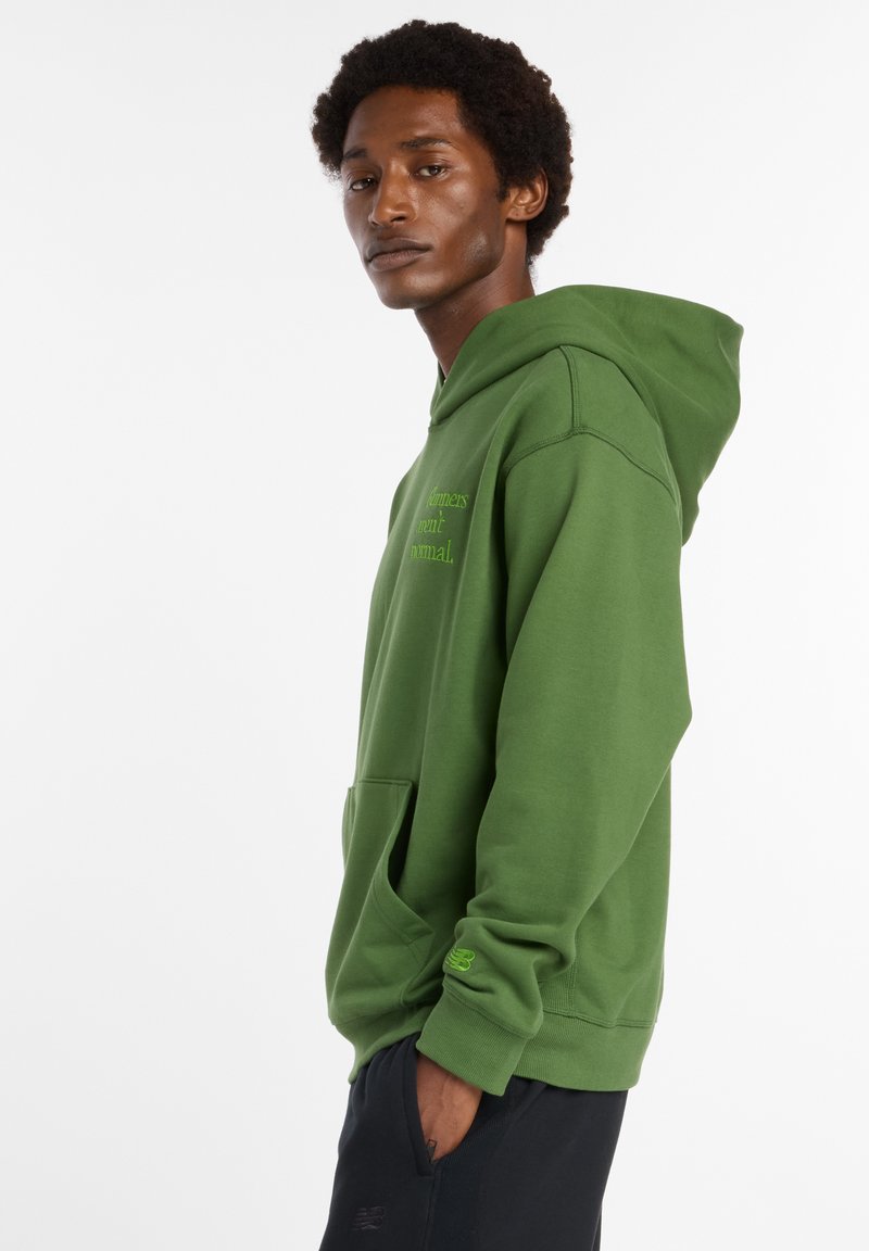 Sweat à capuche vert avec une poche avant, fabriqué en matériau doux. Présente un texte sur la poitrine et un petit logo en vert plus clair sur la manche.