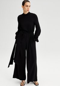 Salopetă neagră cu mâneci lungi, guler înalt, talie ajustată, siluetă cu pantaloni largi și curea legată. Țesătură netedă, ușoară, cu un luciu subtil.
