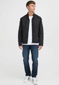 Zwarte jas met een staande kraag, gemaakt van textuurstof. Draag het over een wit t-shirt, gecombineerd met een spijkerbroek en witte sneakers.