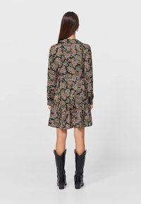 Mujer con cabello largo y lacio vistiendo un vestido de paisley de mangas largas y botas de vaquero negras hasta la mitad de la pantorrilla, de pie de espaldas sobre un fondo liso.