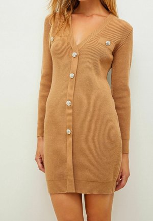 Femme portant une robe cardigan en maille camel à manches longues boutonnée avec des boutons décoratifs argentés et un col en V.