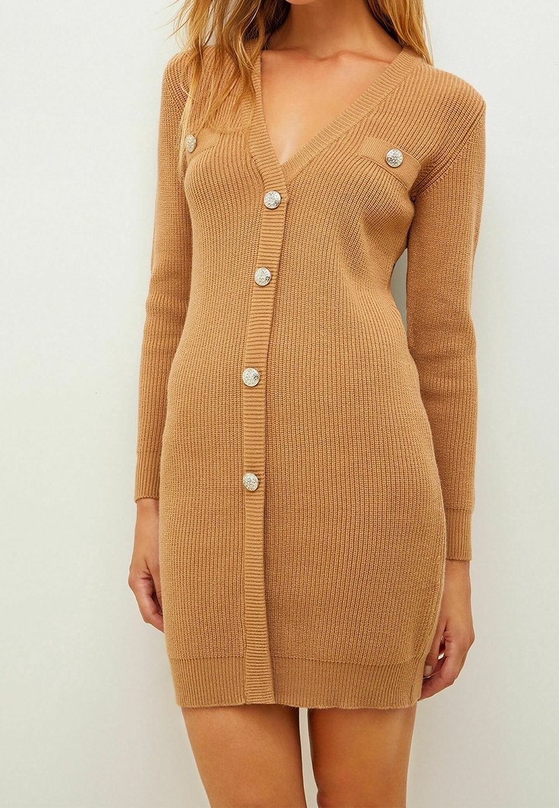 Femme portant une robe cardigan en maille camel à manches longues boutonnée avec des boutons décoratifs argentés et un col en V.