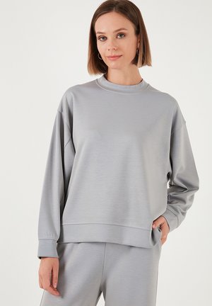 Svijetlosivi dukserica od mekanog materijala, s okruglim izrezom, spuštenim ramenima i rebrastim manžetama. Glatka tekstura s minimalnim dizajnerskim detaljima.
