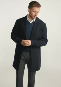 Granatowy wełniany płaszcz z dużymi klapami, jednorzędowy fason i trzy guziki; nałożony na ciemny sweter i dopasowane szare spodnie.