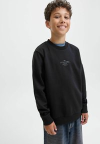 Sudadera negra con cuello redondo, mangas largas y puños acanalados. Presenta un logotipo blanco impreso en el pecho. Usada sobre unos vaqueros.