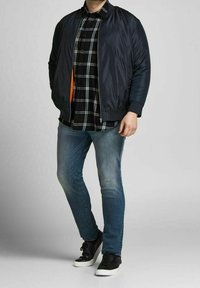 Giubbotto bombardiere navy sopra una camicia a scacchi bianca e nera, abbinato a jeans azzurri chiari e scarpe nere con suole bianche.