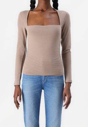 Persoon draagt een nauwsluitend beige top met lange mouwen en een vierkante halslijn, gecombineerd met lichtblauwe spijkerbroek, staand tegen een effen witte achtergrond.