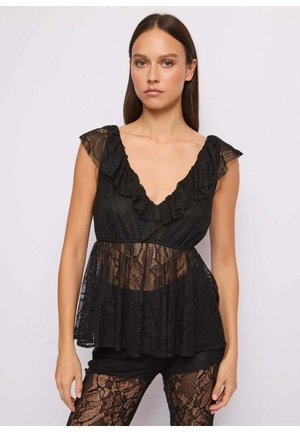 Donna che indossa un top nero senza maniche in pizzo con scollo a V arricciato e leggings trasparenti in pizzo abbinati, in piedi davanti a uno sfondo semplice.