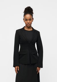 Blazer noir à coupe ajustée, avec des revers crantés, deux poches avant et une fermeture à un bouton. Porté sur une robe noire.
