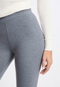 Grijze leggings met een gladde textuur, hoge tailleband en een strakke pasvorm. Een crème-kleurig langmouwen top is gedeeltelijk zichtbaar in het beeld.