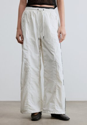 Persoon draagt witte wijde broek met trekkoord in de taille en zwarte schoenen, staand op een grijze vloer tegen een eenvoudige lichte achtergrond.