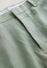 Hellgrüne Leinenhose mit einem flachen Bund und einer subtilen Struktur. Das Innenfutter hat eine gestreifte Fütterung. Sichtbare Nahtdetails entlang der Nähte.