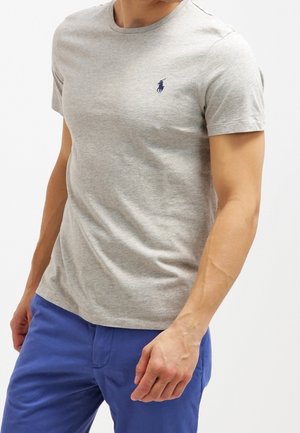 Homme portant un t-shirt à manches courtes gris clair avec un petit logo bleu marine et un pantalon bleu, sur un fond blanc uni.