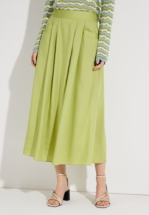 Frau trägt einen hellgrünen Midirock mit Falten und Taschen, kombiniert mit beigen High-Heel-Sandalen und einem blau-grünen Zickzack-Pullover.