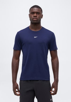 Άνδρας που φοράει μπλε ναυτικό T-shirt Reebok και μαύρα σορτς, στέκεται μπροστά σε ένα απλό ανοιχτόχρωμο φόντο, κοιτάζοντας προς τα εμπρός.