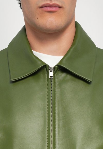 Chaqueta de cuero verde con una textura suave, un cuello amplio y una cremallera en la parte delantera. Incluye una camiseta blanca visible en el escote.