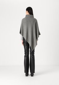 Grå stickad poncho med hög krage, med en draperad design, bärs över svarta läderbyxor. Slät textur och asymmetrisk fåll.