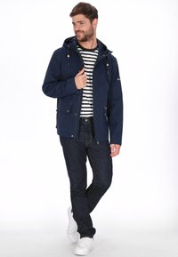 Navyblauwe waterdichte jas met capuchon, ritssluiting aan de voorkant en twee voorzakken. Gecombineerd met een gestreept shirt en donkere jeans, met witte sneakers.